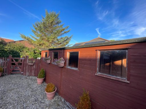 Red wooden shed with windows, surrounded by a gravel pathway and potted plants.