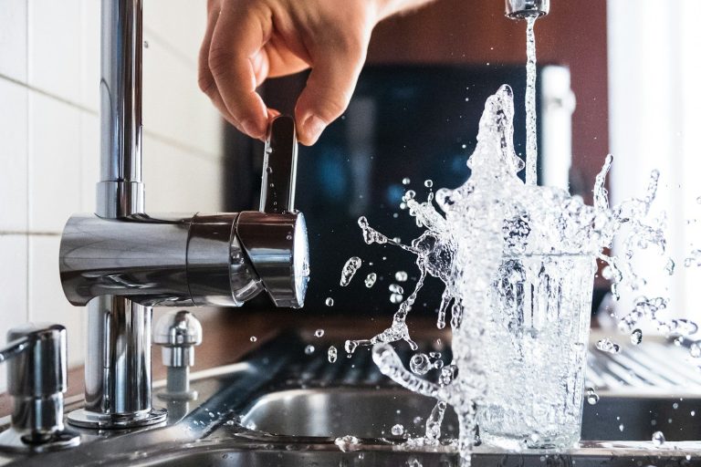 Hand pouring water into a glass, causing a splash at the kitchen sink.