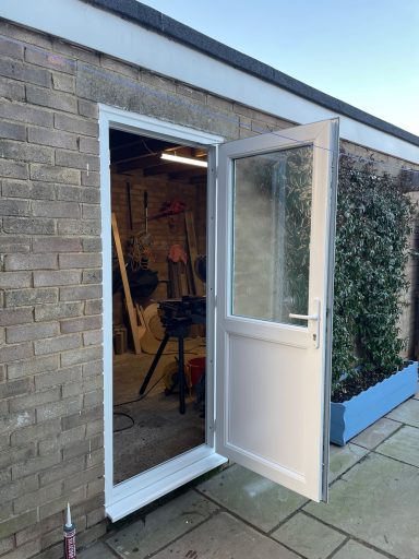 White door standing open, revealing a shed interior with tools and greenery outside.