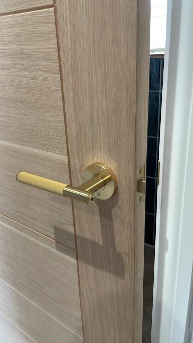 Close-up of a wooden door with a gold handle partially open, revealing a dark tiled area.
