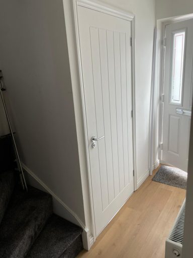 White paneled door next to an open front door in a hallway with wooden flooring.