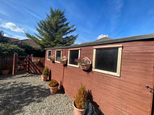 Wooden shed with potted plants and window boxes, set in a gravel area under a blue sky.